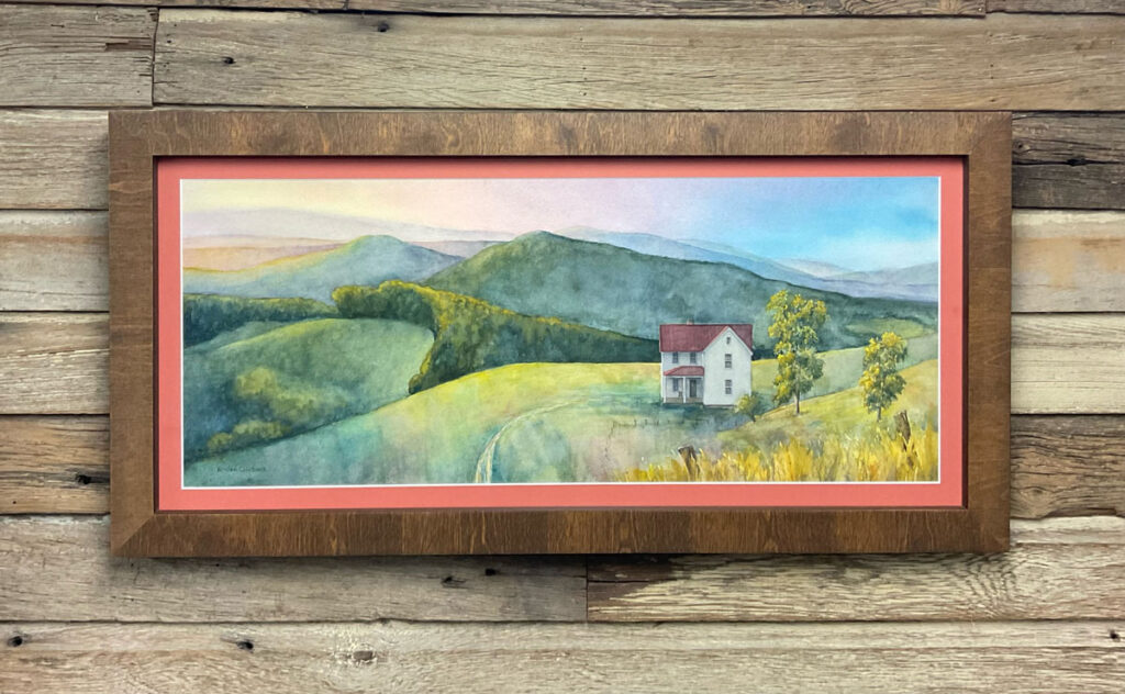 A watercolor in a frame hung on a wall. The watercolor shows rolling hills and a white farmhouse in a field.